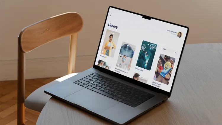 A laptop sits on a table next to a chair. The screen displays an interface with the heading "Library." There are several category cards with titles such as "Medical procedures & protocols" and "Patient safety." In the top right corner, there is a user image with a name.