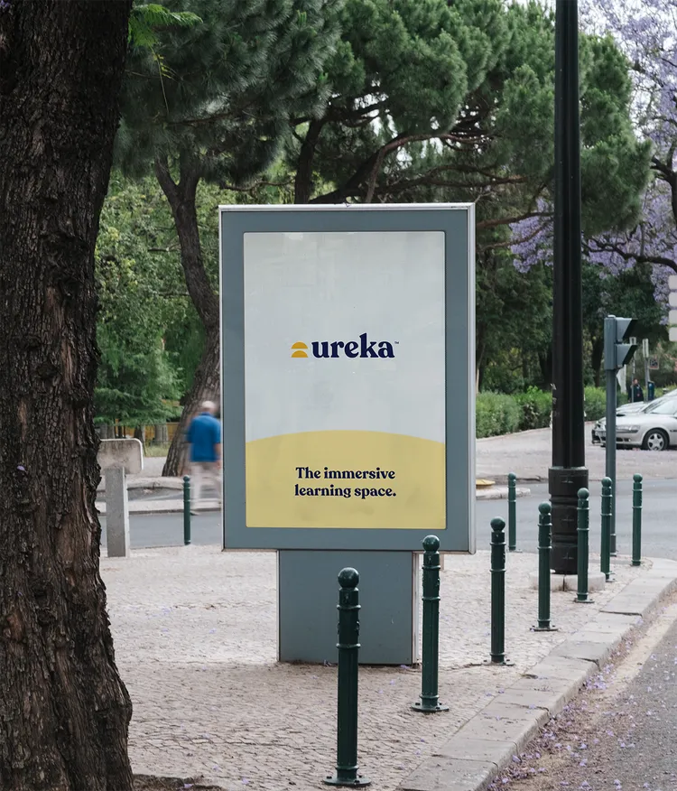 An advertising poster in an urban setting displays the 'Ureka' logo with the slogan 'The immersive learning space'. The poster is on a paved sidewalk, surrounded by trees and parked cars.