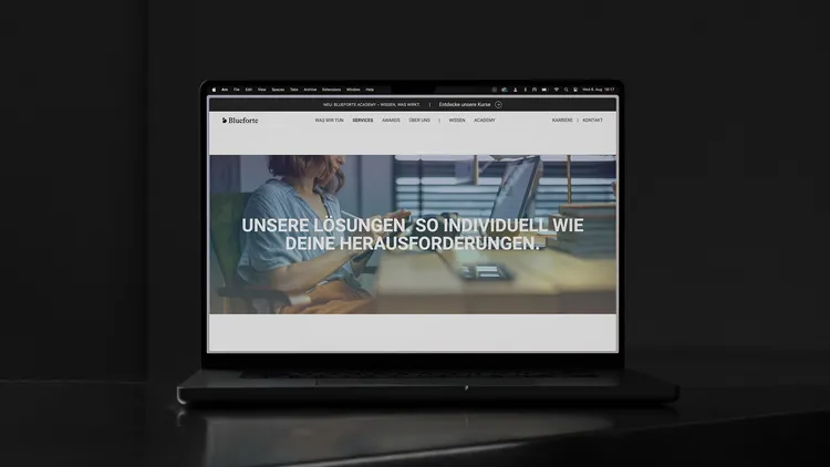 A laptop on a table displays a Blueforte website. The screen shows a person working on a laptop with the text 'OUR SOLUTIONS, AS INDIVIDUAL AS YOUR CHALLENGES.'.