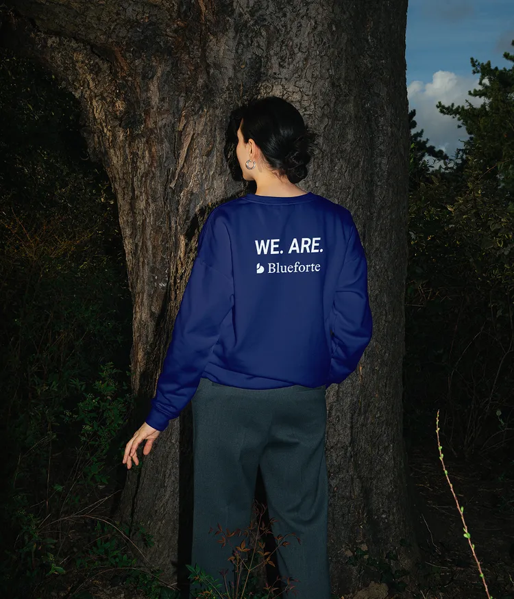 A person stands with their back to the camera in front of a tree. They are wearing a blue top with the white text 'WE. ARE. Blueforte' on the back. There are trees and shrubs around the tree.
