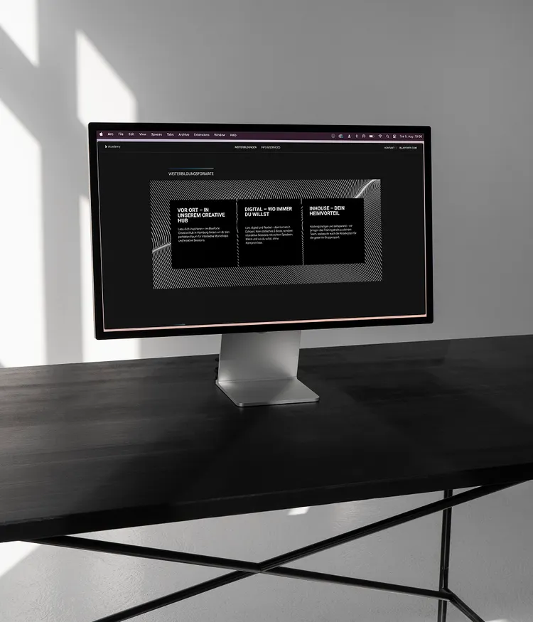 A monitor sits on a black table displaying a website. The website features information about educational formats with headings 'On-site – In our Creative Hub', 'Digital – Wherever You Want', and 'In-house – Your Home Advantage'. The room is minimalistic with shadows on the wall.