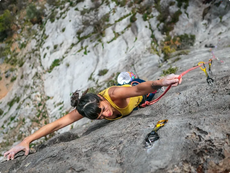 A woman in a yellow top climbs a steep rock face. She is secured and holds onto a rope with one hand while reaching for a hold with the other hand. Her climbing gear is visible, including a harness and carabiners.
