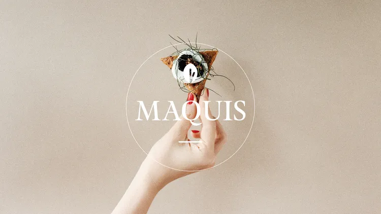 The Maquis logo above a hand presenting an artfully garnished cracker.