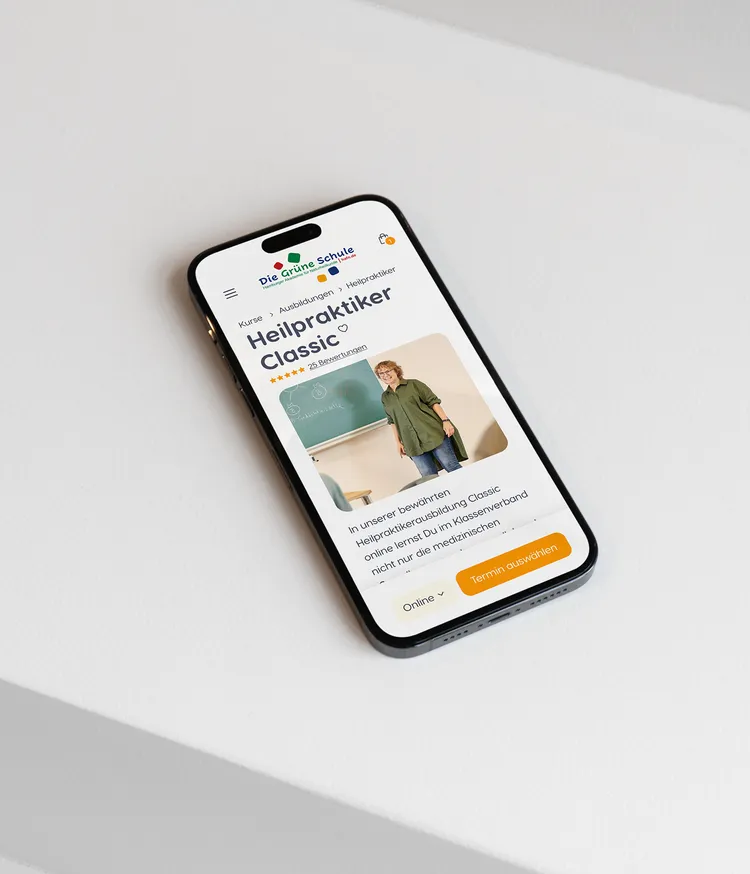 A smartphone is lying on a white table displaying a webpage from 'Grüne Schule', presenting a course called 'Heilpraktiker Classic'. An image of a person standing in front of a blackboard is shown, with an option to select an appointment.