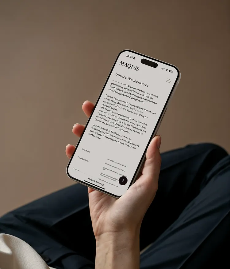 A person is holding a smartphone displaying a restaurant's website called 'MAQUIS.' The screen shows the text 'Unsere Wochenkarte' describing the seasonal and regional dishes. The background is brown and out of focus.