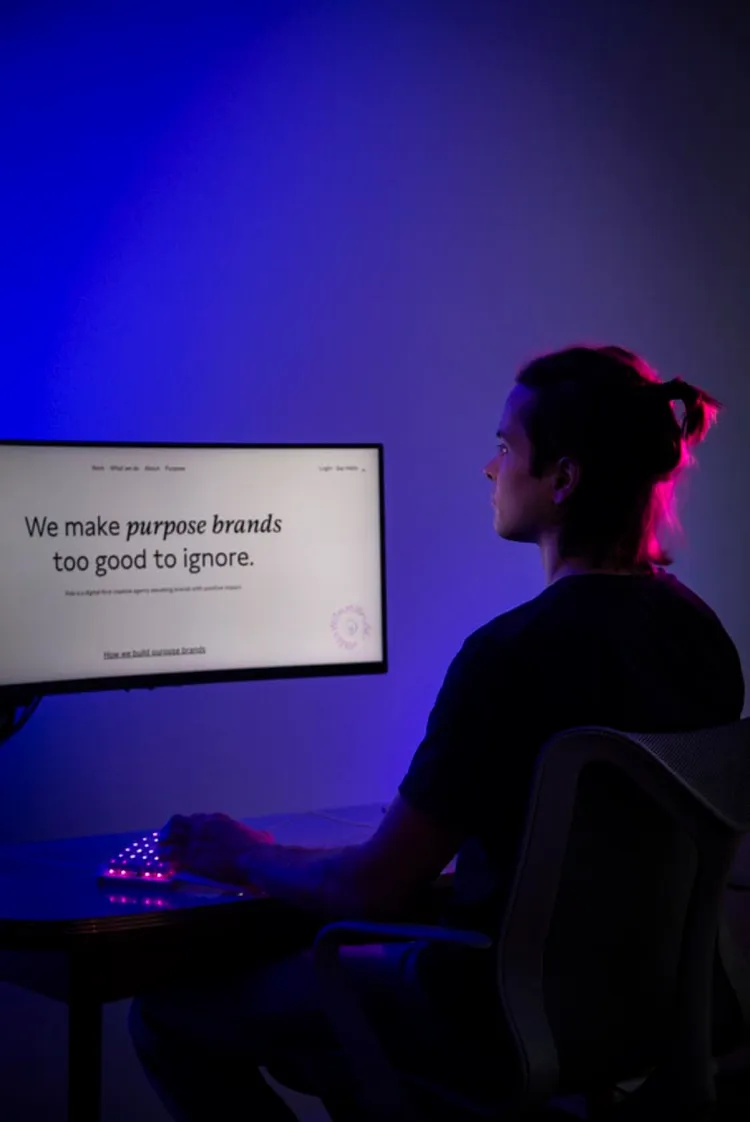Ein Mann mit einem Dutt sitzt vor einem Computerbildschirm, der eine Website mit dem Text 'We make purpose brands too good to ignore' zeigt. Der Raum ist in blauen und pinken Farben beleuchtet, und die Tastatur hat bunte Lichter.