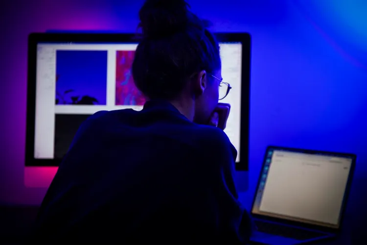 Person sitzt vor zwei Bildschirmen, beide mit blauer Beleuchtung. Der große Bildschirm zeigt bunte Grafiken, während auf dem Laptop-Display ein einfaches Textdokument geöffnet ist. Die Person stützt den Kopf auf die Hand und arbeitet konzentriert.