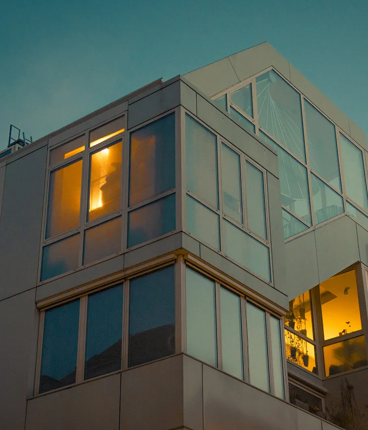A modern building with large glass windows, through which warm yellow light shines. The sky in the background is blue. Plants are visible in one of the windows.