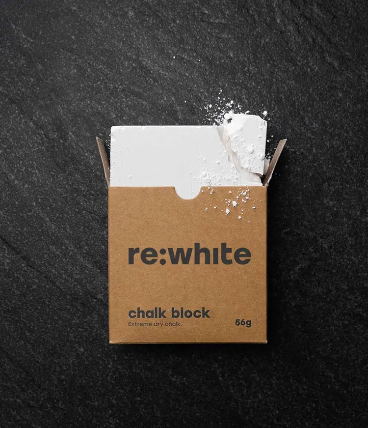 A brown cardboard box with the text "re:white chalk block" and "Extreme dry chalk. 56g" is placed on a dark surface. A white chalk block protrudes from the open box, slightly crumbled, with chalk powder visible on the box.
