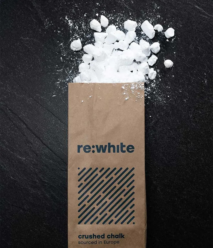 A brown paper bag labeled 're:white crushed chalk sourced in Europe' lies on a dark surface. The bag is open with white, crushed chalk pieces spilling out.