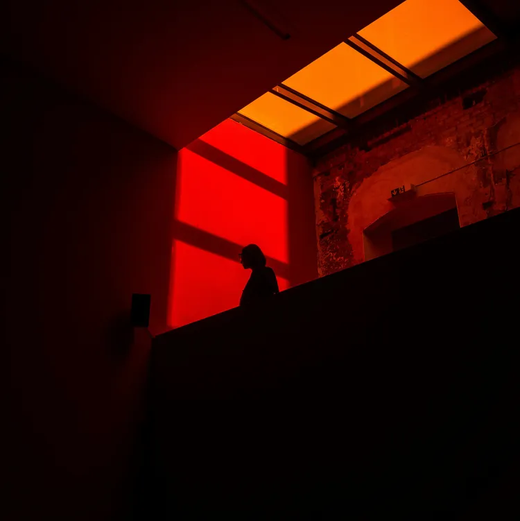 Eine Silhouette einer Person steht vor einem Fenster in einem Raum mit roten und orangen Lichteffekten. Der Raum hat eine Backsteinwand und eine Deckenöffnung, die gelbes Licht hereinlässt.