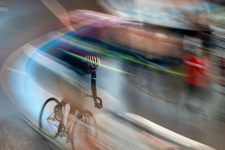A cyclist with a striped helmet rides on a racetrack. The image has motion blur, conveying speed, and the background is blurred.