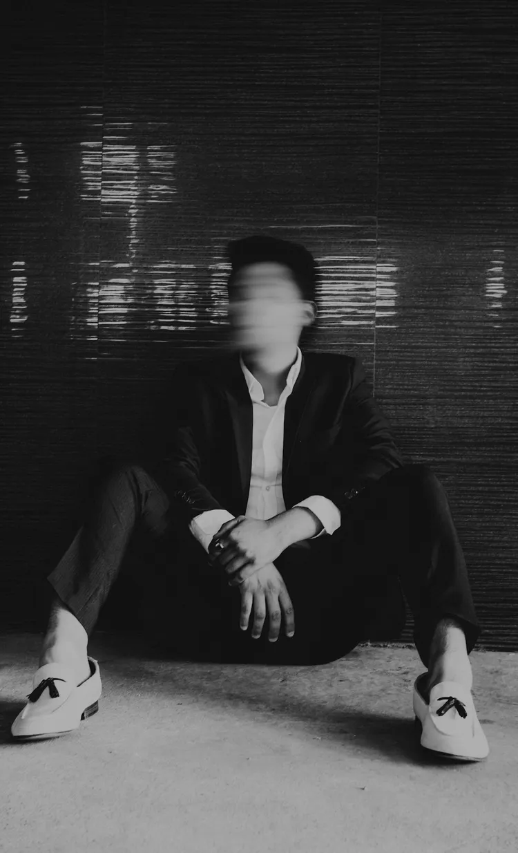 Black and white photo of a person in a suit sitting against a dark wall with a blurred face. The person is wearing white tassel shoes.