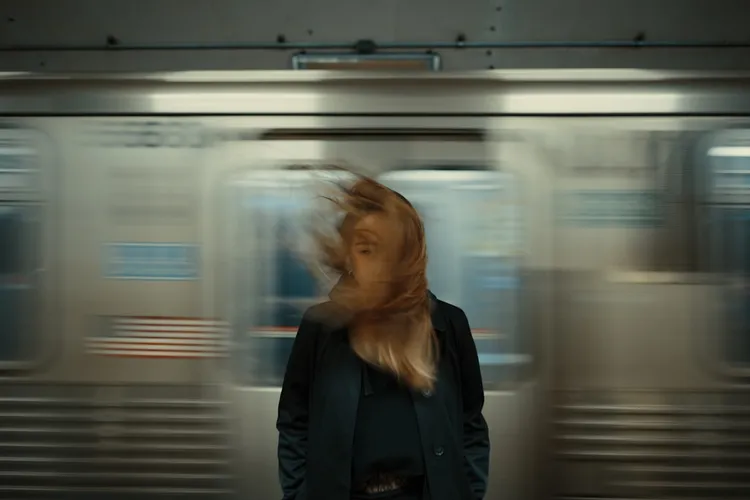A person stands in front of a passing subway train. Their hair is blown by the wind, partially covering their face. The train and the surroundings are motion blurred.