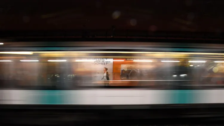 A blurred commuter train speeds past a brightly lit platform with posters. In the background, people and advertisements are faintly visible.